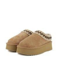 Zoccoli in suede marrone con suola piatta beige, con un accento intrecciato attorno al colletto e una finitura morbida e texturizzata.