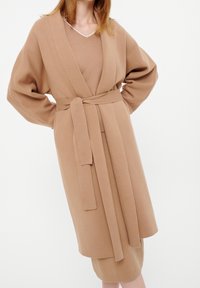 Beige-värinen vyöllinen neuletakki leveillä hihoilla, valmistettu pehmeästä kankaasta. Siinä on syvä V-pääntie ja sen alla on samaa neulosta oleva mekko.