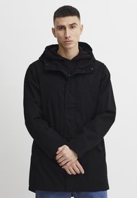 Sort jakke med hætte, der har en frontlynlås og trykknapper, struktureret stof og en let oversize pasform. Stylet over en sort hættetrøje.