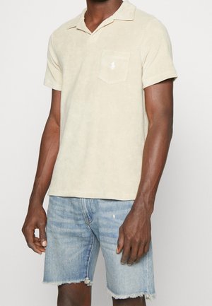 Mężczyzna w beżowej krótkiej koszulce polo z logo na kieszeni i postrzępionych jasnoniebieskich szortach dżinsowych, stoi na białym tle.