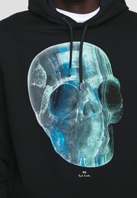 Černá mikina s kapucí s velkým průsvitným motivem lebky v modrých tónech, s hladkou texturou. Šňůrky v tmavě zelené barvě s kovovými hroty.