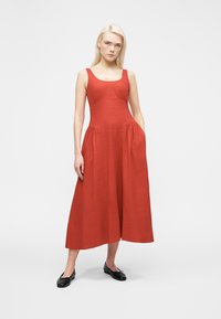 Jeune femme debout portant une robe midi sans manches de couleur rouille avec des poches et des ballerines noires sur un fond blanc uni.