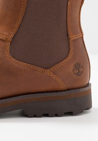 Bottines en cuir marron avec des côtés élastiques, textures contrastées et une semelle en caoutchouc robuste. Présente un logo d'arbre au niveau du talon.