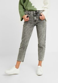 Jeans grises de talle alto con un lavado desgastado, pierna estrecha, dobladillo deshilachado y rayas laterales negras, combinados con un suéter de punto verde y blanco.