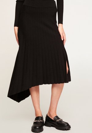 Zwarte geribbelde gebreide midi-rok met asymmetrische zoom en zijsplit, gecombineerd met zwarte chunky loafers met zilveren kettinkdetail.