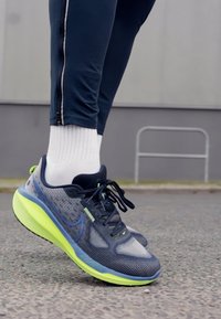 Chaussure de course avec une tige en mesh gris, des accents verts et une semelle rembourrée. Chaussettes blanches simples visibles au-dessus de la cheville.