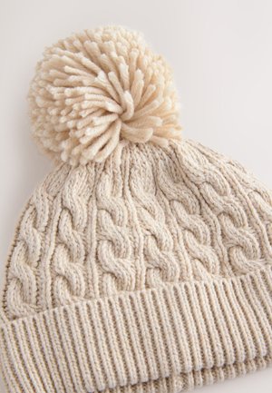 Kötött bézs sapka texturált fonott mintával, nagy pomponnal a tetején és bordázott szegéllyel alul.