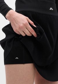 Χέρι σηκώνει μαύρη πλισέ φούστα για να αποκαλύψει εφαρμοστά μαύρα σορτς με μικρό λευκό λογότυπο στον μηρό.