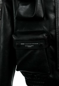 Chaqueta de cuero negra con un bolsillo frontal destacado, banda elástica en la cintura y detalles con la marca en texto plateado.