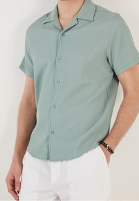 Koszula z krótkim rękawem w jasnym zielonym kolorze, z prążkowaną teksturą i klasycznym kołnierzykiem. Materiał wydaje się lekki i miękki.