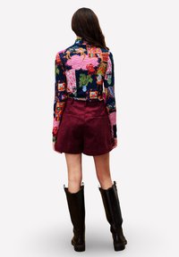 Frau, die ein bunt gemustertes Langarmoberteil, burgunderfarbene Shorts und hohe schwarze Stiefel trägt, mit dem Rücken zur Ansicht stehend.