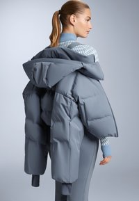 Puhasti sivi kaput s velikom kapuljačom, sa debelim kvadratnim dizajnom. Nosite preko plavog džempera s uzorkom, prikazujući slojevite teksture.