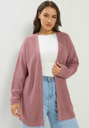 Ung kvinde iført en pink strikket åben cardigan over en hvid skjorte og lyseblå højttaljede jeans mod en hvid baggrund.