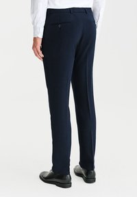 Granatowe spodnie o dopasowanym kroju, z jedną kieszenią z tyłu i subtelną teksturą. Noszone z białą koszulą i czarnymi butami.