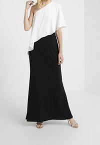 Kobieta ubrana w bezrękawową czarną sukienkę maxi z asymetrycznym białym nakryciem na jednym ramieniu oraz srebrnymi wysokimi sandałami.