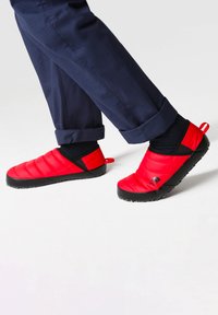 Scarpe slip-on rosse isolate con un design trapuntato, suole in gomma nera e un piccolo logo. Indossate con pantaloni blu scuro a risvolto e calze nere.