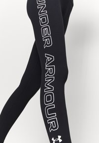 Leggings neri realizzati in tessuto elasticizzato, con un grande logo bianco "UNDER ARMOUR" sul lato e un piccolo logo vicino alla caviglia.