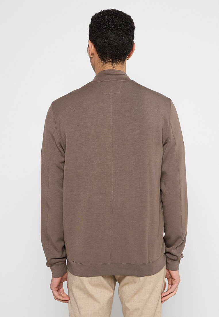 Mężczyzna w brązowej koszulce z długim rękawem i beżowych spodniach stoi tyłem do jednolitego białego tła.