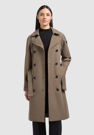 Ženska, ki nosi bež dvoredni trenčkot čez črni puli in črne hlače, stoji z eno roko v žepu plašča.