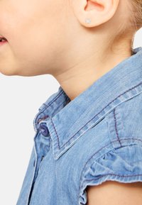 Denimhemd mit kragenförmigem Design, blauer Farbe und lila Naht; Kind trägt einen kleinen Schmetterlingsohrstecker im Ohr.