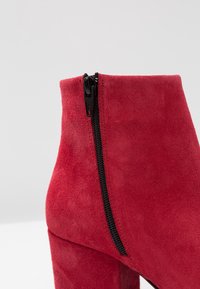 Bottine en daim rouge avec un détail de fermeture éclair noire sur le côté et une forme élégante et structurée. Surface texturée qui ajoute de l'intérêt visuel.