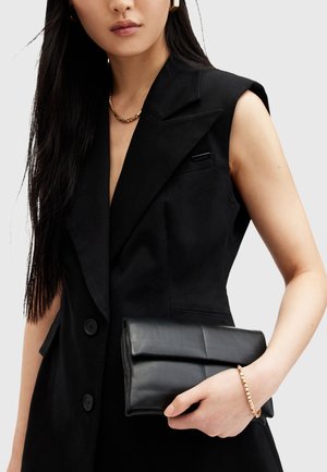 Femme portant un blazer noir sans manches, tenant une pochette noire en cuir pliée, avec un collier chaîne en or et un bracelet clouté en or.