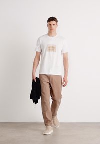 Hvit t-skjorte med trykt design på brystet, kombinert med beige bukser og lette sneakers. Modellen holder en svart jakke.