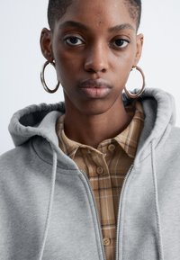 Grau zip-up Kapuzenpulli mit Fleece-Textur, getragen über einem braunen Karo-Hemd mit Knöpfen. Besitzt eine große Kapuze und auffällige silberne Ohrreifen.