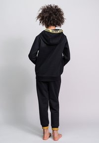Zwarte hoodie en sweatpants set met een gele "BATMAN" logo op de kraag. De broek heeft gele manchetten bij de enkels.