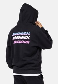 Felpa nera con tasca a marsupio, ampia cappuccio e logo "ROSSIGNOL" colorato in gradiente pastello sul retro. Polsini e orlo a coste.