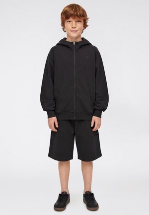 Junge mit hellbraunen Haaren, der eine schwarze Zip-up-Kapuze, schwarze knielange Shorts und schwarze Sneakers trägt, steht vor einem schlichten grauen Hintergrund.