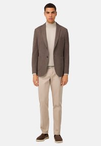 Brun texturerad blazer över en ljus polotröja, parat med beige byxor och bruna slip-on skor. Enkel, skräddarsydd design med fickor.