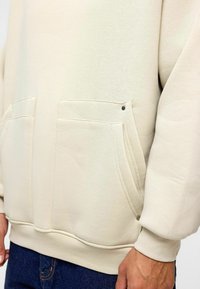 Lichtbeige sweatshirt met een ontspannen pasvorm, voorzien van twee voorzakken, zichtbare stiksels en een subtiele metalen accent op één zak.
