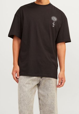 Homme portant un t-shirt noir oversize avec un graphisme blanc de soleil, lune et cercle sur la poitrine gauche, associé à un jean gris clair.