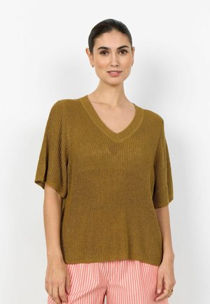 Frau mit zurückgebundenem dunklem Haar, die ein lockeres, olivgrünes Strickoberteil mit V-Ausschnitt und rosa-weiß gestreifte Hose trägt, vor weißem Hintergrund stehend.