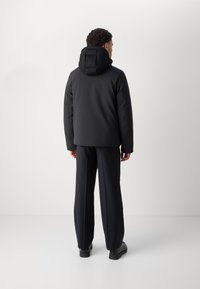 Black insulated jacket with a high collar and textured hood, paired with relaxed-fit black trousers and black shoes, viewed from behind.