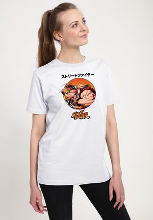 Jonge vrouw met paardenstaart die een wit T-shirt draagt met Street Fighter-personages Ryu en Ken in een vechtpose, met Japanse tekst erboven.