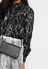 Czarna bluzka z abstrakcyjnym wzorem z białych linii, z falbankowanym kołnierzem, zestawiona z szarą spódnicą w wężowy wzór oraz małą czarną skórzaną torebką na ramię.