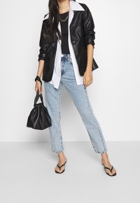 Svart läderblazer över en vit skjorta med knappar, tillsammans med ljusblå rakbenjeans och svarta flip-flops. Svart dragsko-väska hålls i handen.