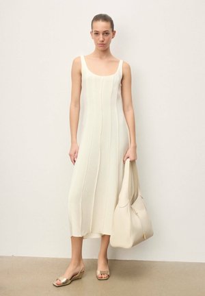 Mujer con un vestido midi blanco sin mangas, sosteniendo un bolso grande de color marfil y usando sandalias doradas de cuña con punta abierta sobre un fondo liso.