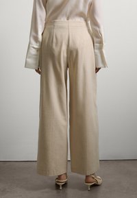 Personne portant un pantalon beige taille haute et large, un blouse crème à manches longues avec des poignets allongés, et des escarpins beiges à bride arrière, se tenant sur un sol en béton.