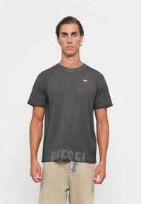 Jeune homme avec des cheveux courts et ondulés portant un t-shirt noir délavé de Diesel et un pantalon beige clair, se tenant devant un fond blanc uni.