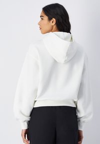 Haut à capuche blanc au fit décontracté, avec capuche et poignets côtelés. Matière douce avec une texture lisse, porté avec un pantalon noir, design minimal.