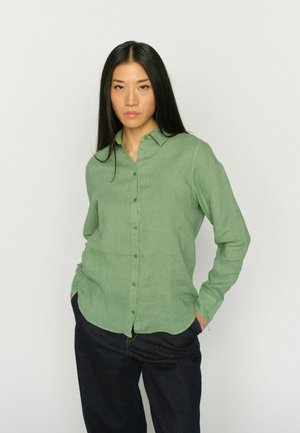 Žena s dlhými čiernymi vlasmi, oblečená v zelenej košeli so zapínaním na gombíky a tmavých džínsoch, stojaca s rukami v vreckách na neutrálnom pozadí.