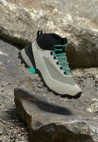 Bottes de randonnée légères de couleur beige avec des accents noirs et des lacets turquoise. Elles disposent d'une semelle texturée et d'une conception imperméable. Logo affiché en évidence.