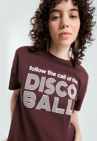 Bordeauxfarbenes Baumwoll-T-Shirt mit einem auffälligen weißen Grafiktext-Design, das "folgen Sie dem Ruf der DISCO BALL" in verschiedenen gestreiften Schriftarten zeigt.
