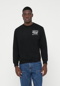 Junger Mann in einem schwarzen Sweatshirt mit einem weißen Auto-Graphic und Text auf der Brust, kombiniert mit blauen Jeans, vor einem weißen Hintergrund stehend.