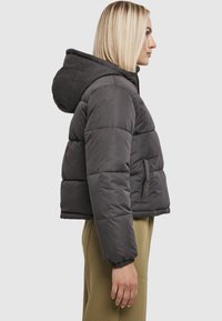 Mørkegrå dunjakke med hætte, der har quiltet design, elastiske manchetter og sidelommer. Båret over lysegrønne bukser.