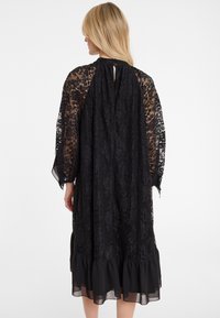 Schwarzes Spitzenkleid mit transparenten Ärmeln, einem Schlüsselloch-Rückenteil und einem rüschengesäumten Saum. Der Stoff hat ein florales Muster und ist leicht.