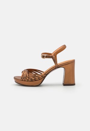 Sandale à talons hauts métallisée brune avec un large talon bloc, présentant un design de brides croisées, une bride de cheville réglable et une texture lisse.